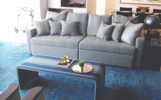 sala de estar, living, com tapete azul textura, mesa de centro em tons azuis listrados, par de cadeiras brancas com acabamento em madeira nos braços. sofá 4 lugares, cinza com almofadas.